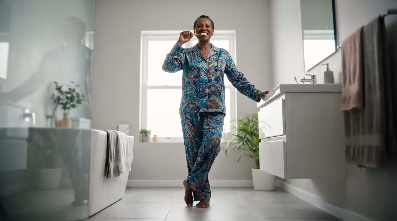 An older woman performs a single-leg balance exercise while brushing her teeth in a sunlit bathroom.